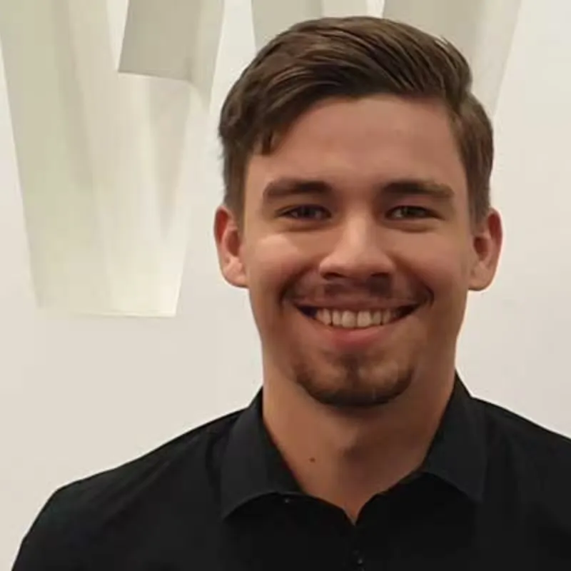 Ein junger man in einem schwarzen Oberteil steht vor einem weißen Rewe Logo