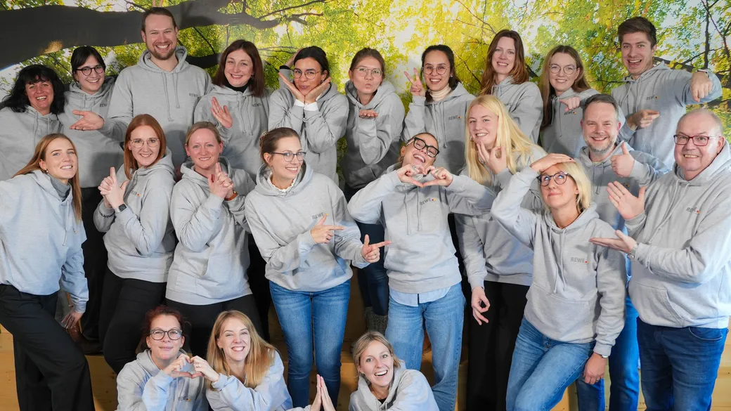 Mehrere Personen stehen vor einer grünen Wand und tragen graue Rewe Pullover.
