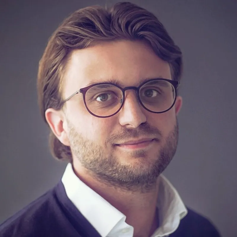 Ein Portrait von einem Mann mit einer runde Brille, er traegt einen Anzug