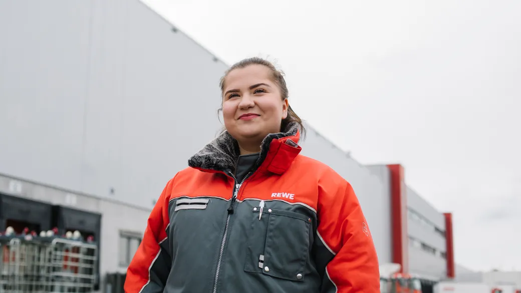 Ein Frau steht in einer roten REWE Jacke vor einer Logistik Halle. 